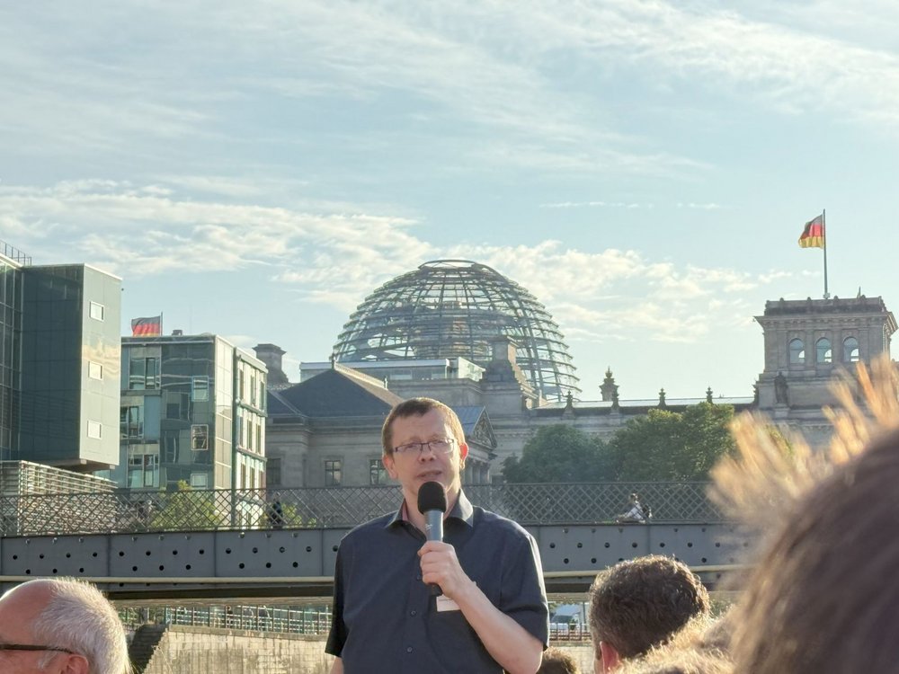 Ein Referent hält eine Ansprache über ein Mikrofon auf dem Oberdeck eines Schiffes, während im Hintergrund die gläserne Kuppel des Reichstagsgebäudes in Berlin zu sehen ist.