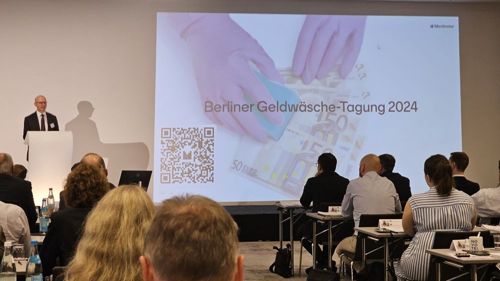 Ein Referent hält einen Fachvortrag am Rednerpult der Berliner Geldwäsche-Tagung vor einer großen Projektionsfläche.